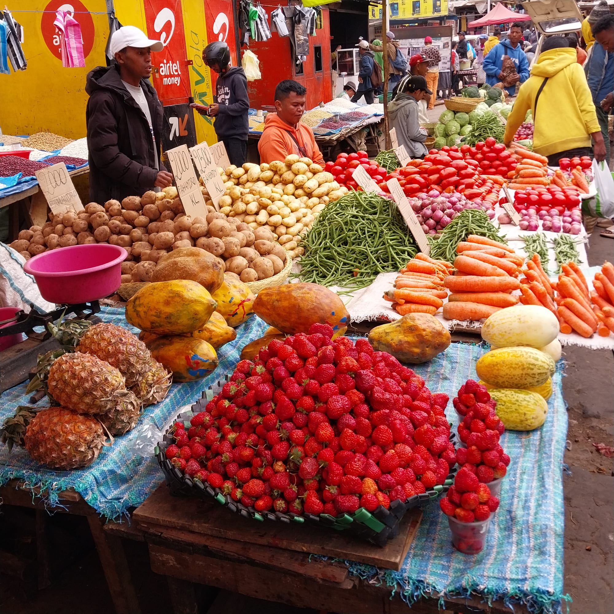 A market stall piled high with potatoes, carrots, tomatoes, strawberries and melons, with vendors and shoppers behind it
