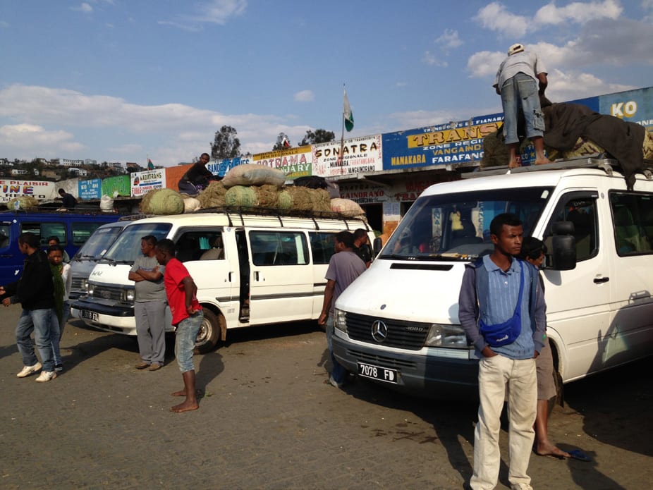 Several white minibuses loaded with sacks on their roof racks at a taxi-brousse station, with passengers and drivers standing around them