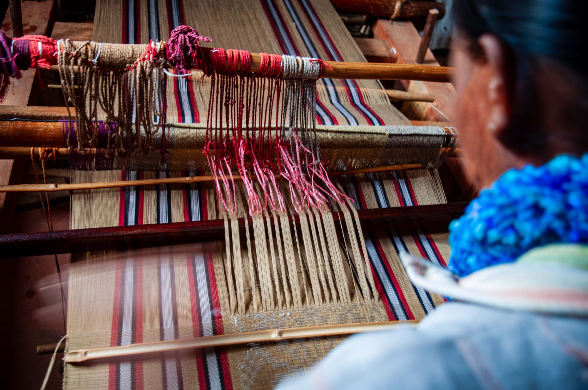 A woman seen from behind works at a wooden loom threaded with fine silk in natural, red and dark blue stripes