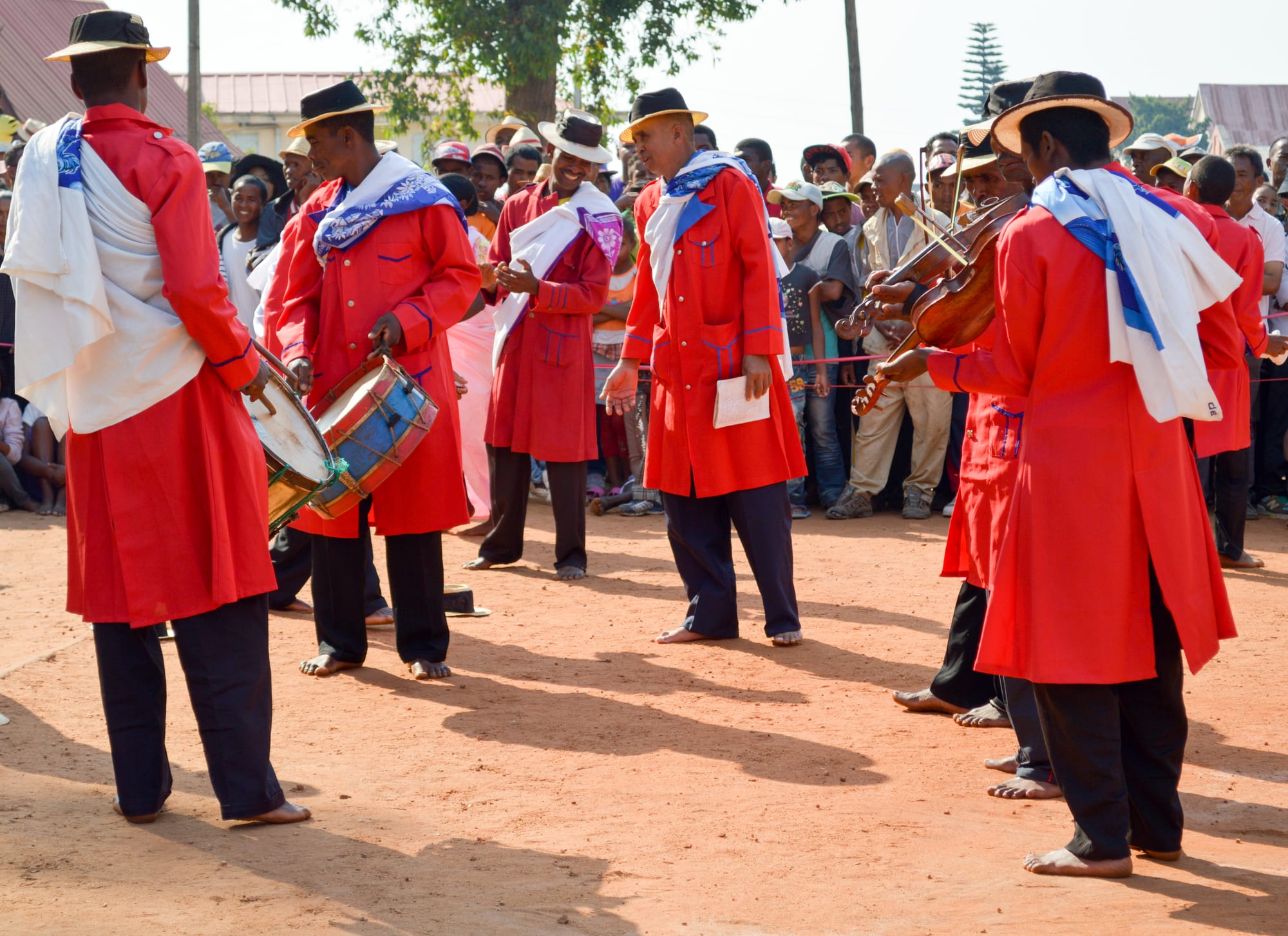 A troupe of hira gasy performers in bright red frock coats, white shawls and dark straw hats plays drums and violins barefoot on open red earth, watched by a crowd behind a rope