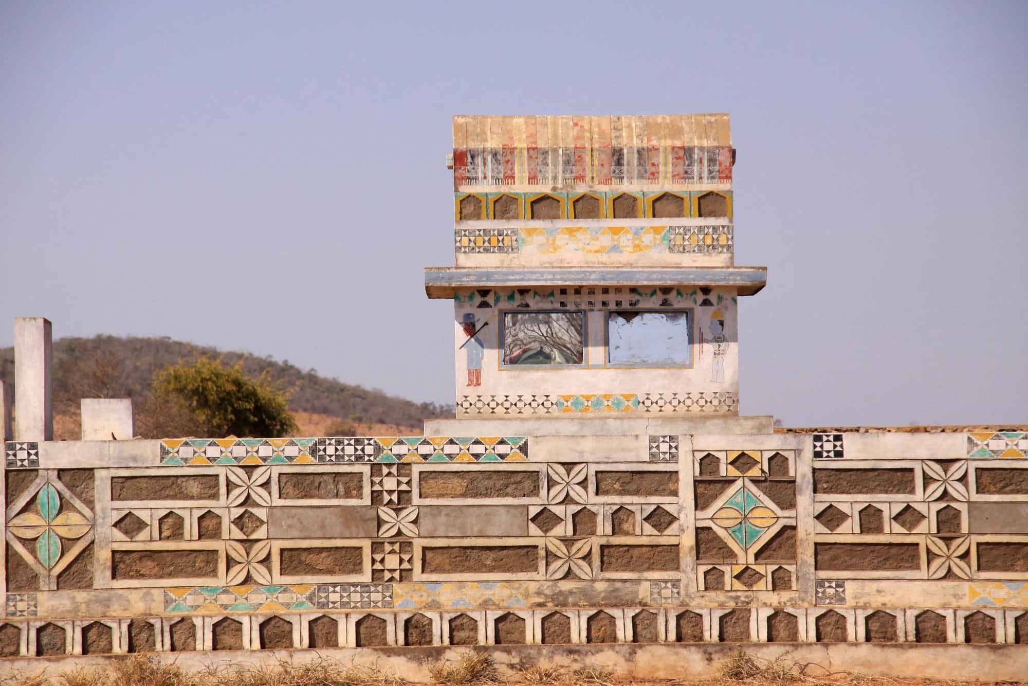 A long stone tomb wall decorated with painted geometric panels in blue, yellow and green, with a small painted tower rising at its centre against a clear sky