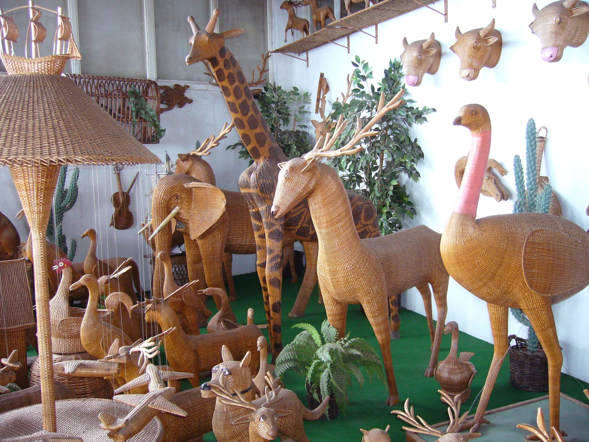 Life-size wicker animals including a giraffe, deer, elephant and ostrich stand crowded together in a showroom, with wicker animal heads mounted on the wall above