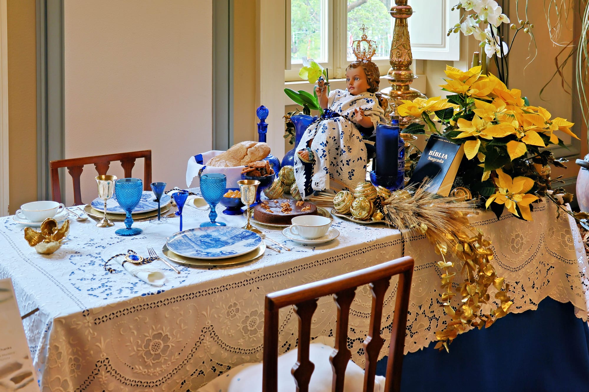 A table laid for Christmas with a white cutwork embroidered tablecloth, blue glassware, blue and white plates, bread, a cake, yellow poinsettias and a crowned figure of the infant Jesus