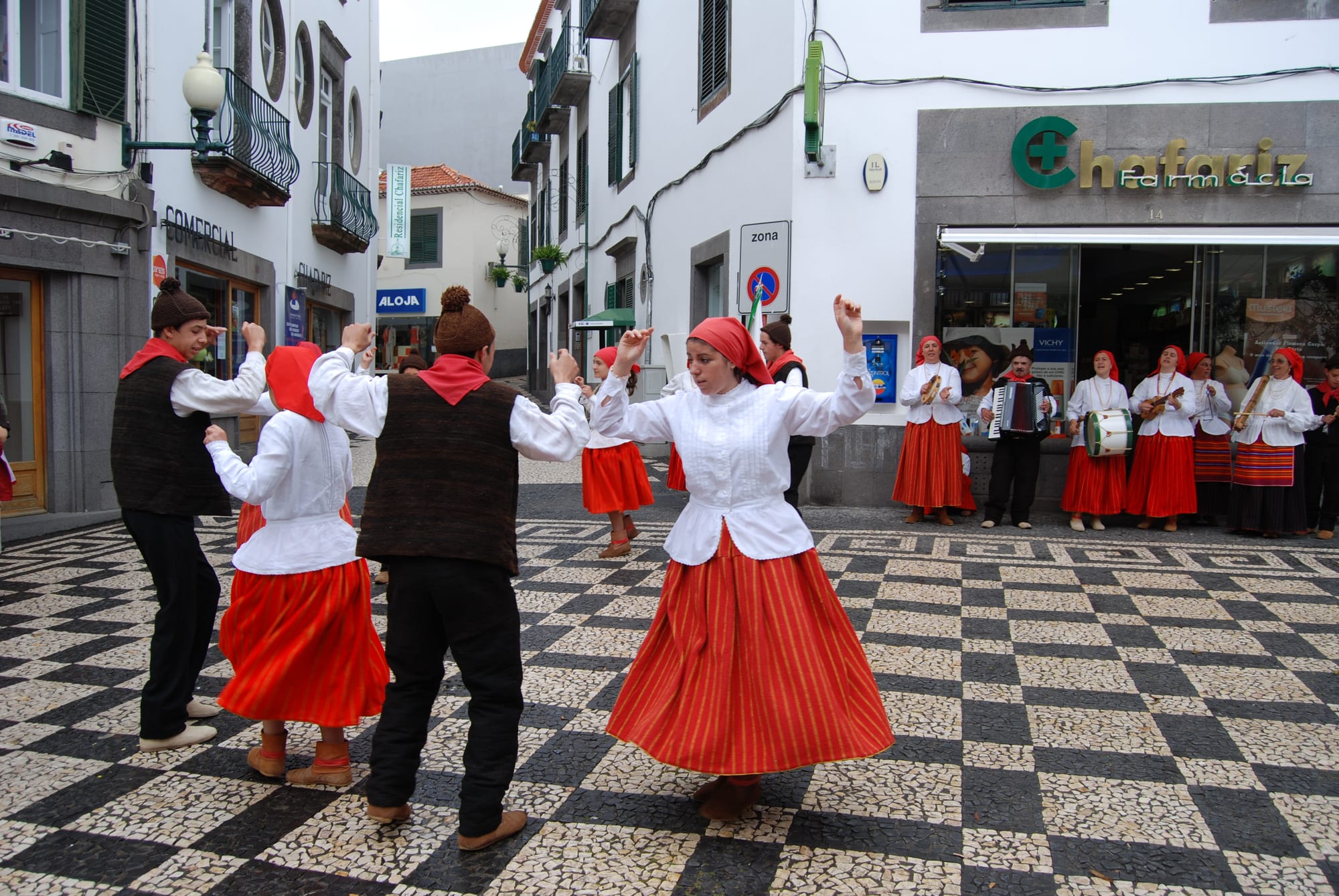 Dancers in red and orange striped skirts, white blouses and red headscarves dance on a black and white cobbled street, with musicians in matching costume playing an accordion, a drum and small guitars behind them