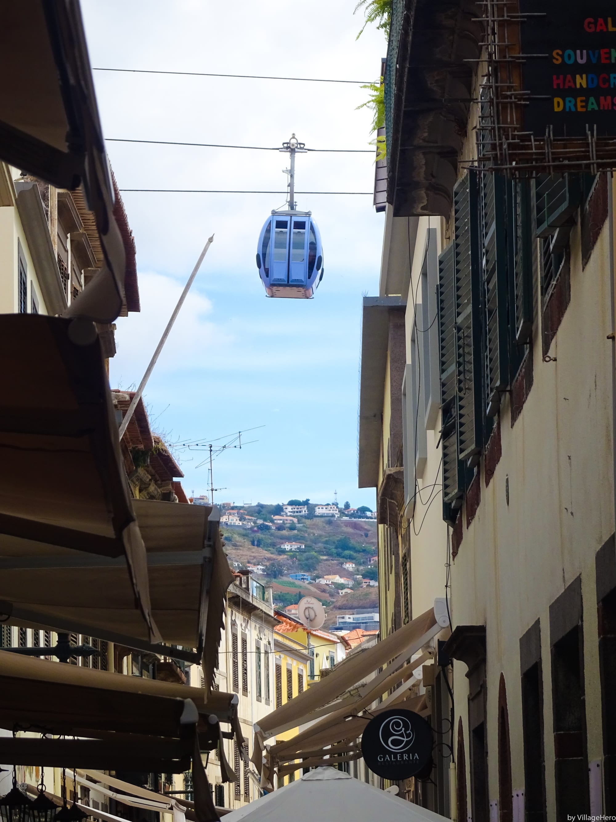 A cable car gondola passing overhead between old town buildings, hillside visible beyond