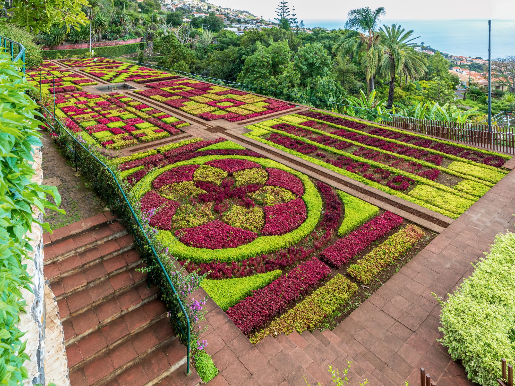 A formal terraced garden with colourful geometric flower beds overlooking a bay