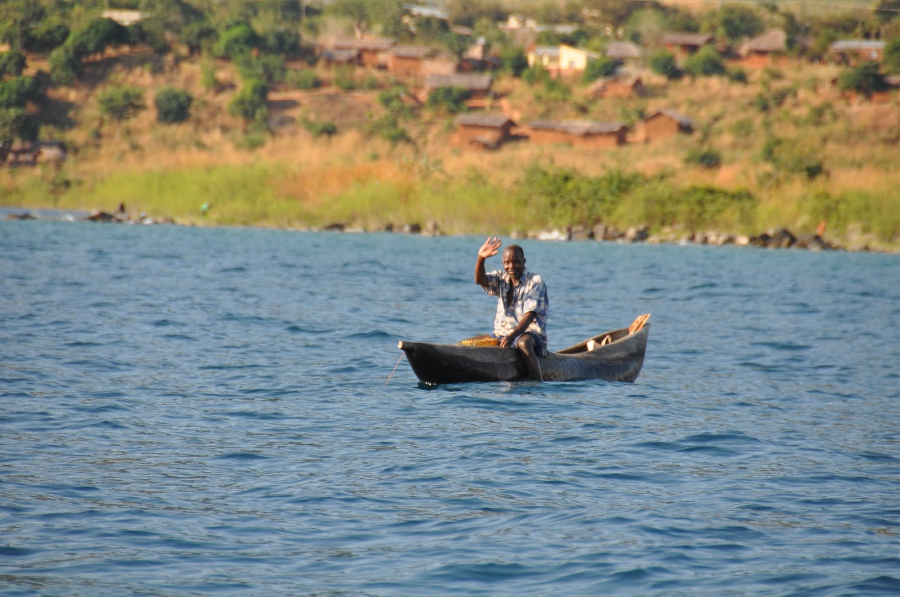 A fisherman waves from a dugout canoe on Lake Malawi, a lakeside village behind
