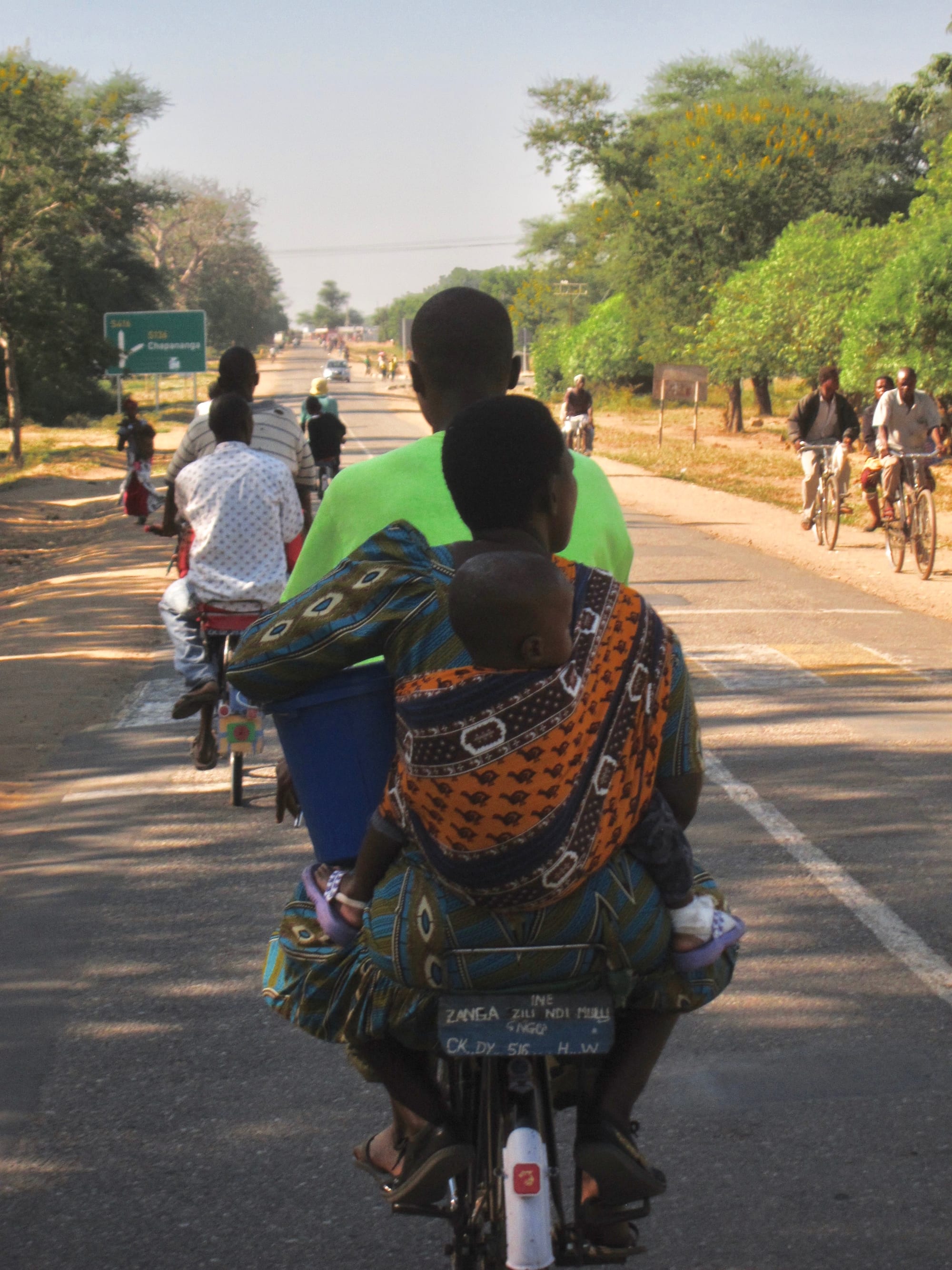 A woman carrying a child on her back rides a bicycle along a tree-lined rural road, with several other cyclists visible ahead and behind