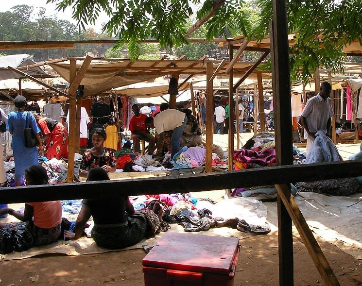 Vendors and shoppers among market stalls piled with clothing and fabric under wooden and tarpaulin awnings