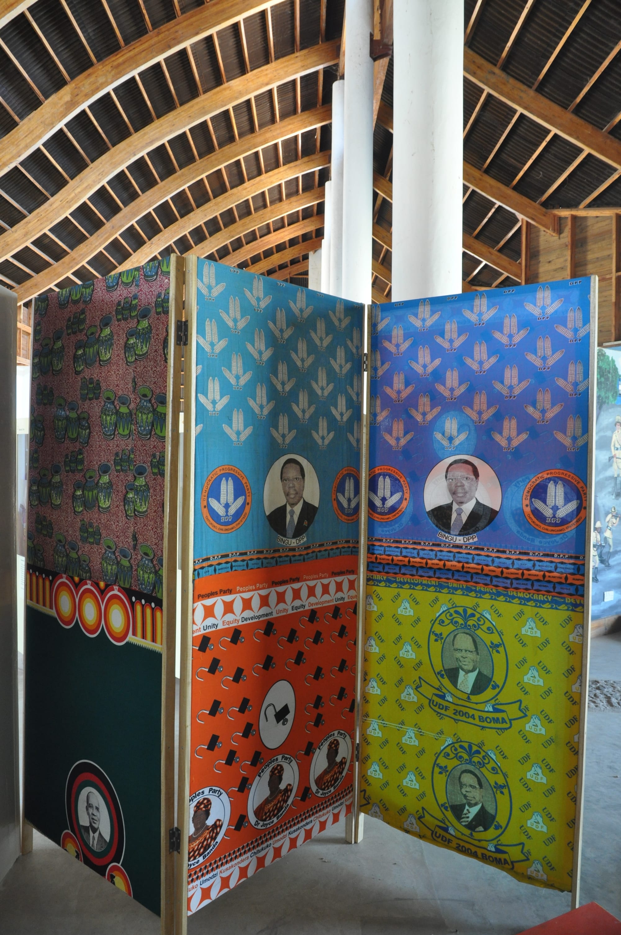 A folding screen covered in brightly printed cloths in blue, orange, yellow and green, bearing political party emblems and portraits of Malawian politicians, standing beneath a curved timber roof