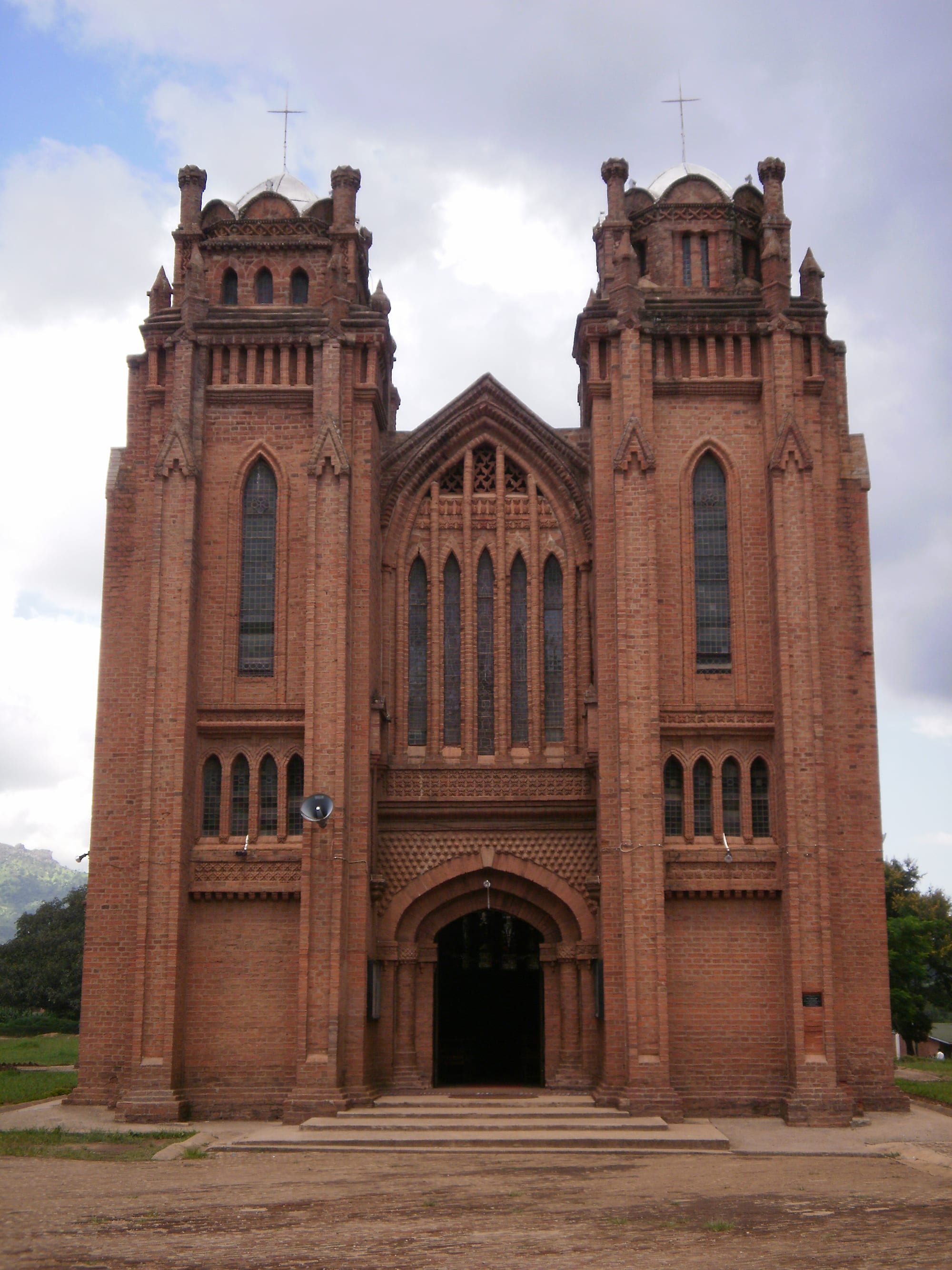 The red-brick facade of a large church with two tall towers topped by small domes and crosses, rows of arched windows and a central arched doorway at the top of a few steps