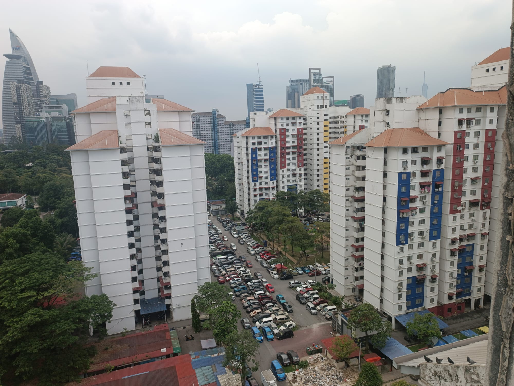A cluster of tall, colour-coded public housing tower blocks in Kuala Lumpur, with cars parked in the courtyard below and the city skyline behind