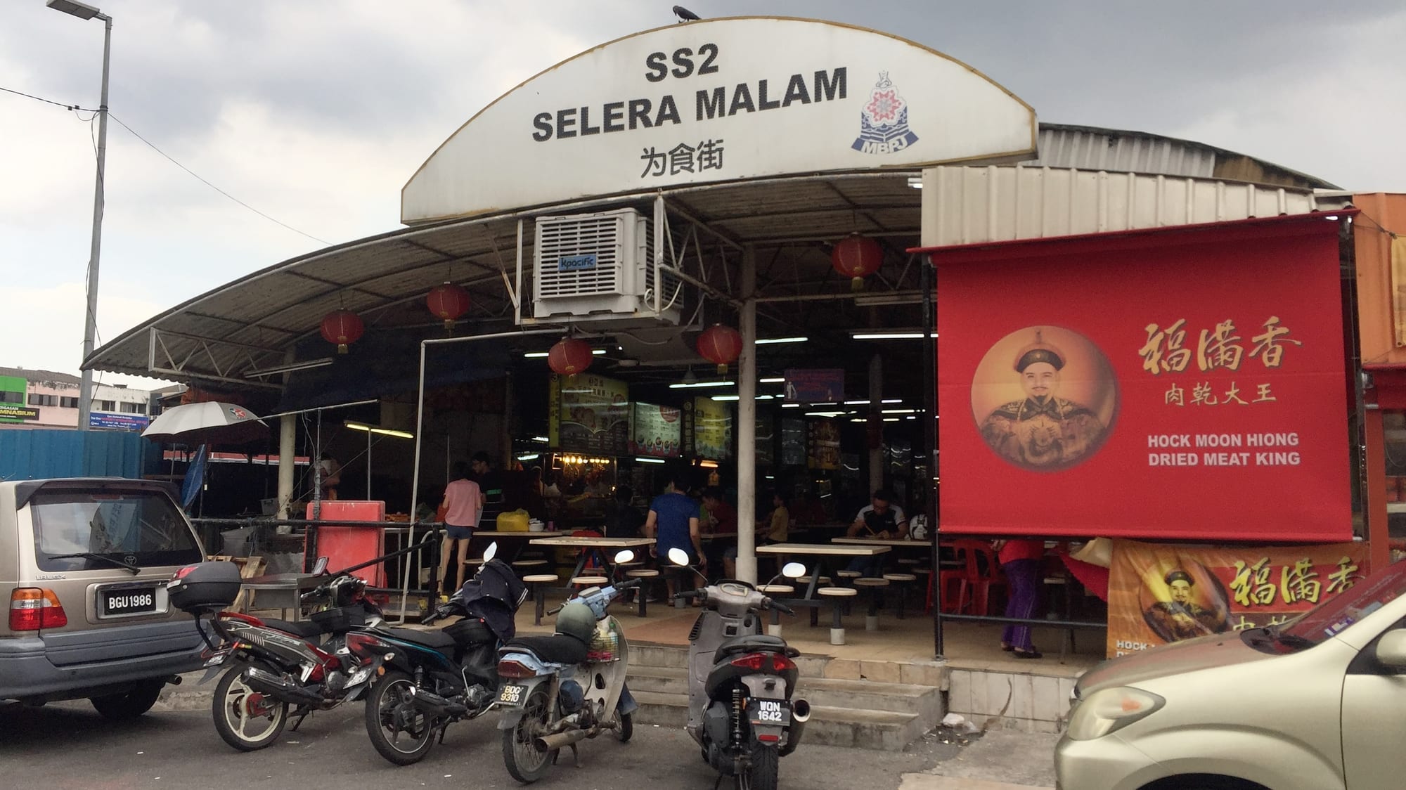 An open-sided hawker centre with red lanterns, food stalls, diners seated at tables, and motorcycles parked outside
