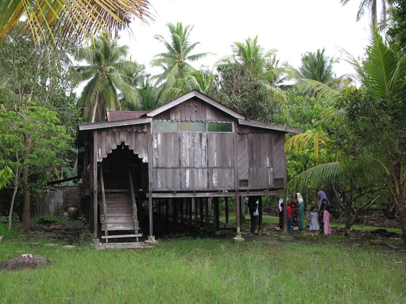 A traditional wooden stilt house in a Malaysian village, surrounded by palm trees, with a family standing beside it