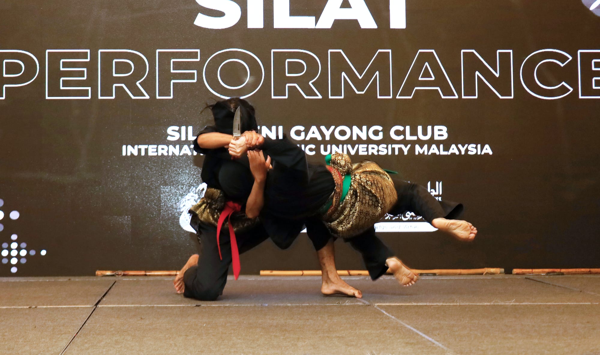 Two silat practitioners in black with gold and red sashes grapple on a stage, one flinging the other, who holds a knife, sideways through the air