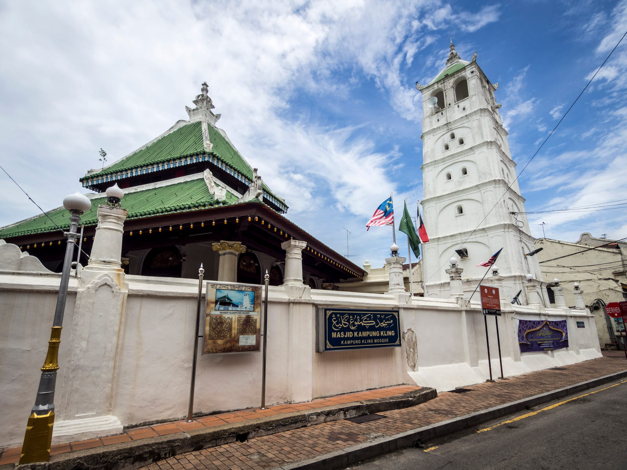A mosque with a tiered green pyramid roof behind a white perimeter wall, beside a tall white minaret with arched openings and a small green dome, under a blue sky with light cloud