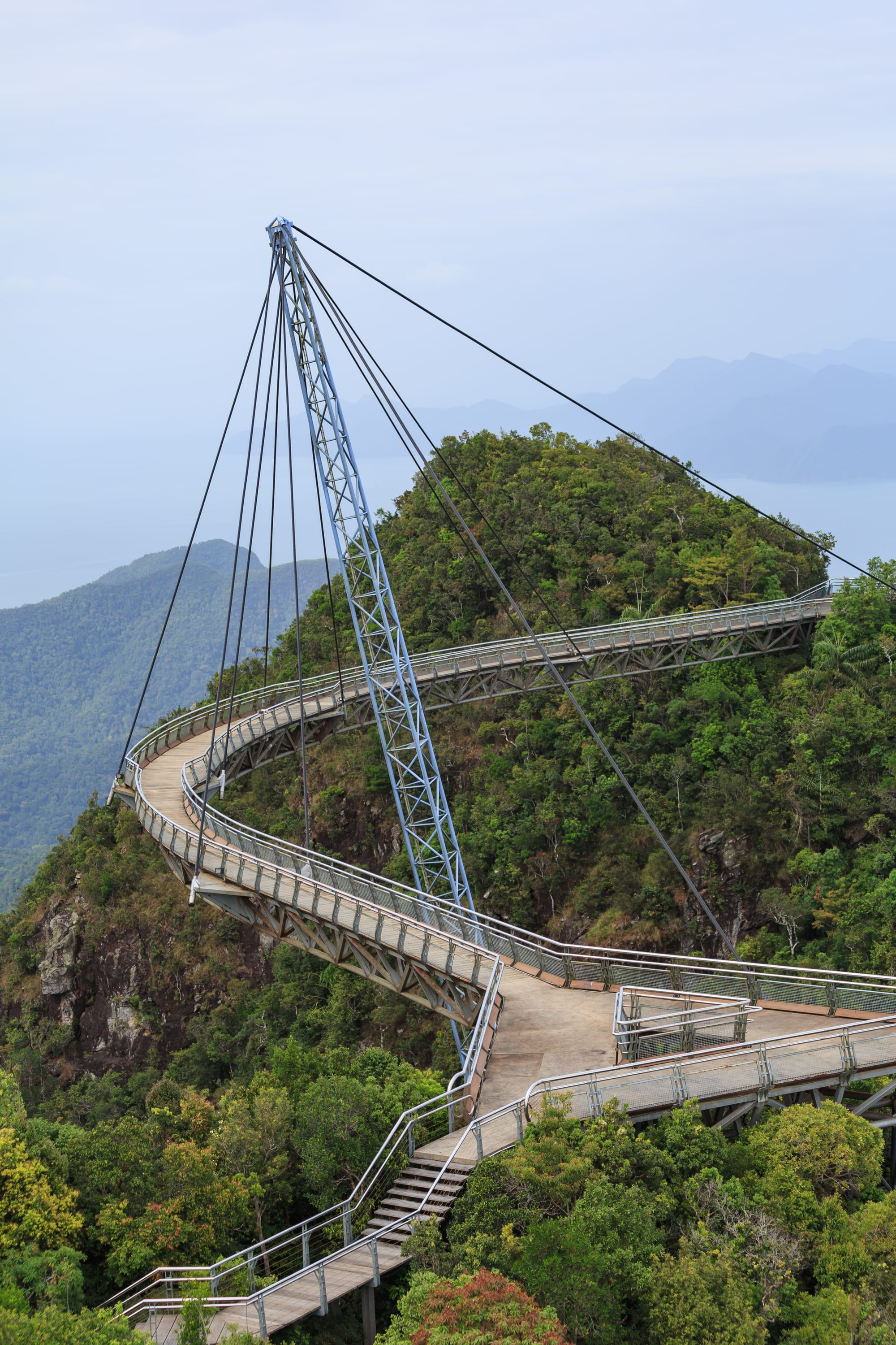 A curved pedestrian bridge suspended high above forested mountains and the sea