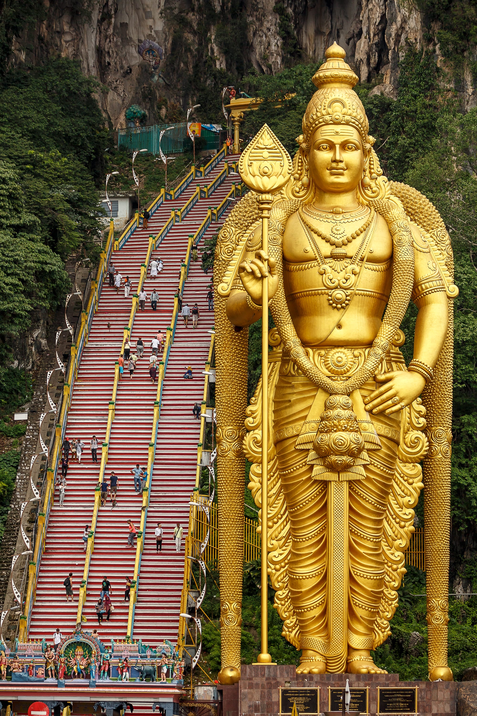 A towering golden statue of a Hindu deity beside a steep flight of colourful steps leading into a limestone cave