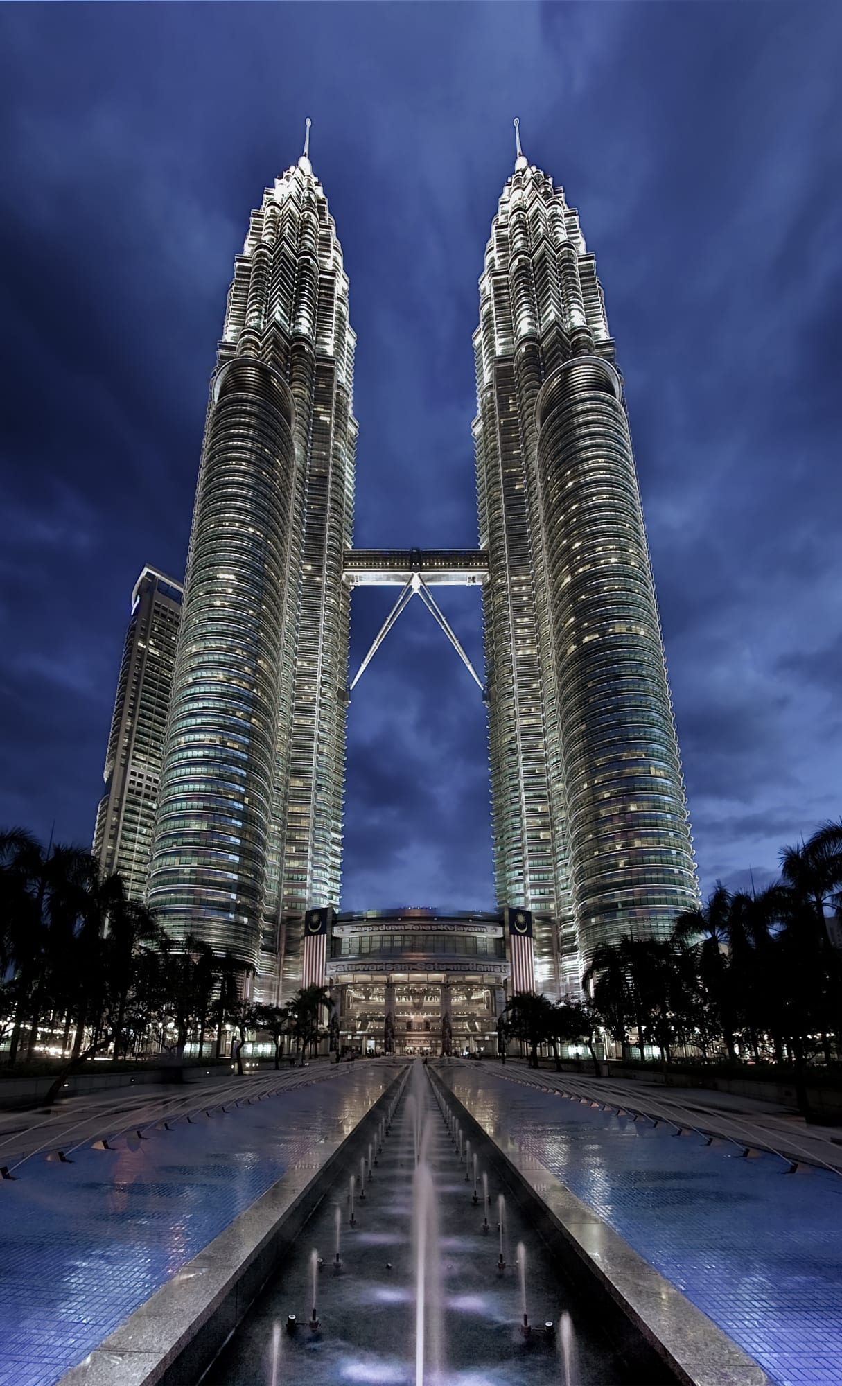 Two identical, ornately detailed silver skyscrapers linked by a sky bridge, lit at dusk