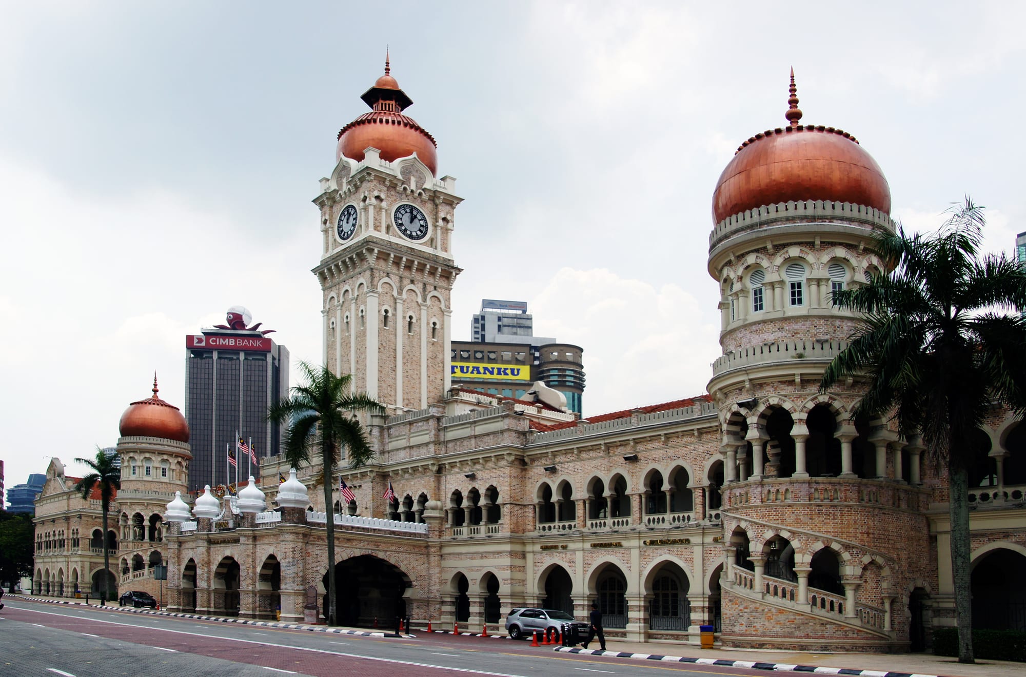 A long brick colonial building with a central clock tower and copper-coloured domes, fronted by palm trees
