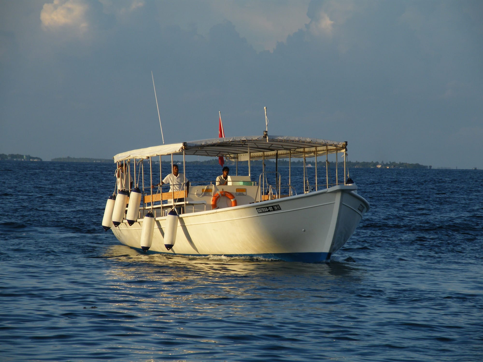 A traditional Maldivian dhoni boat with a canopy roof, carrying several crew members, moving through calm water at sunset with an island visible in the distance