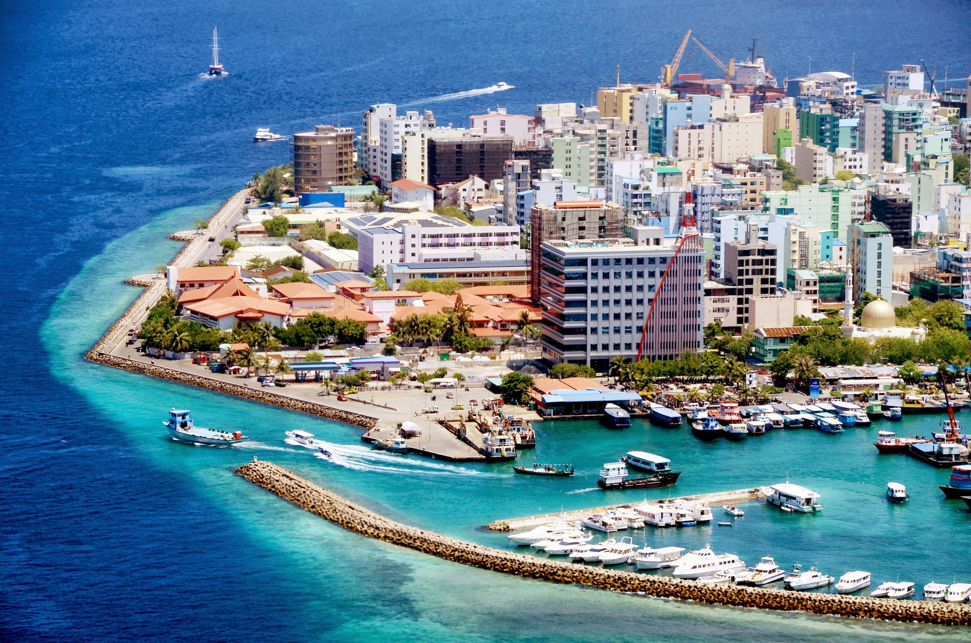 An aerial view of the densely packed high-rise buildings of Malé, ringed by a reef and turquoise water, with boats moored along the harbour