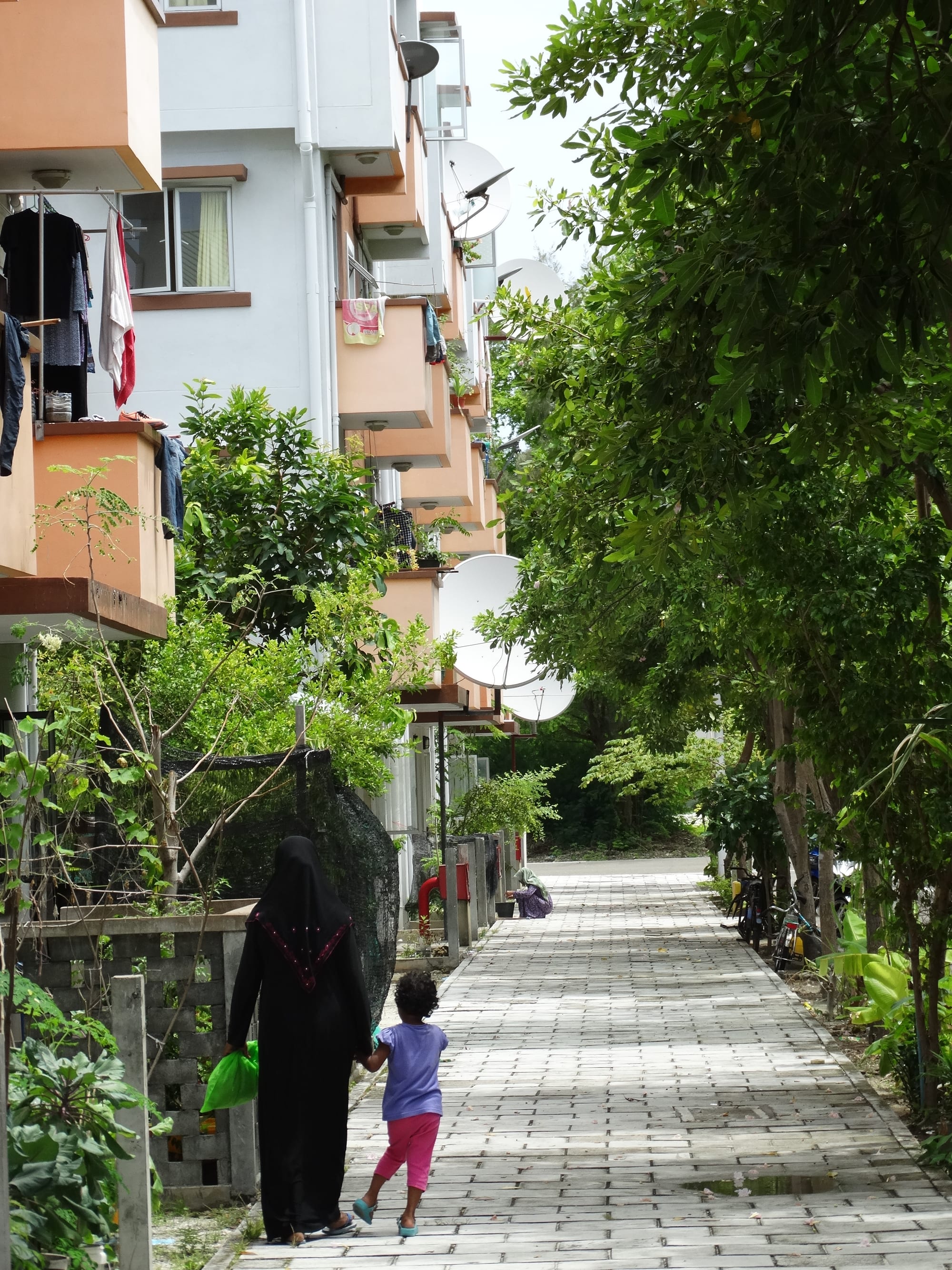 A woman in a black abaya and a young child walk hand in hand along a paved footpath between rows of apartment blocks with laundry hanging from the balconies