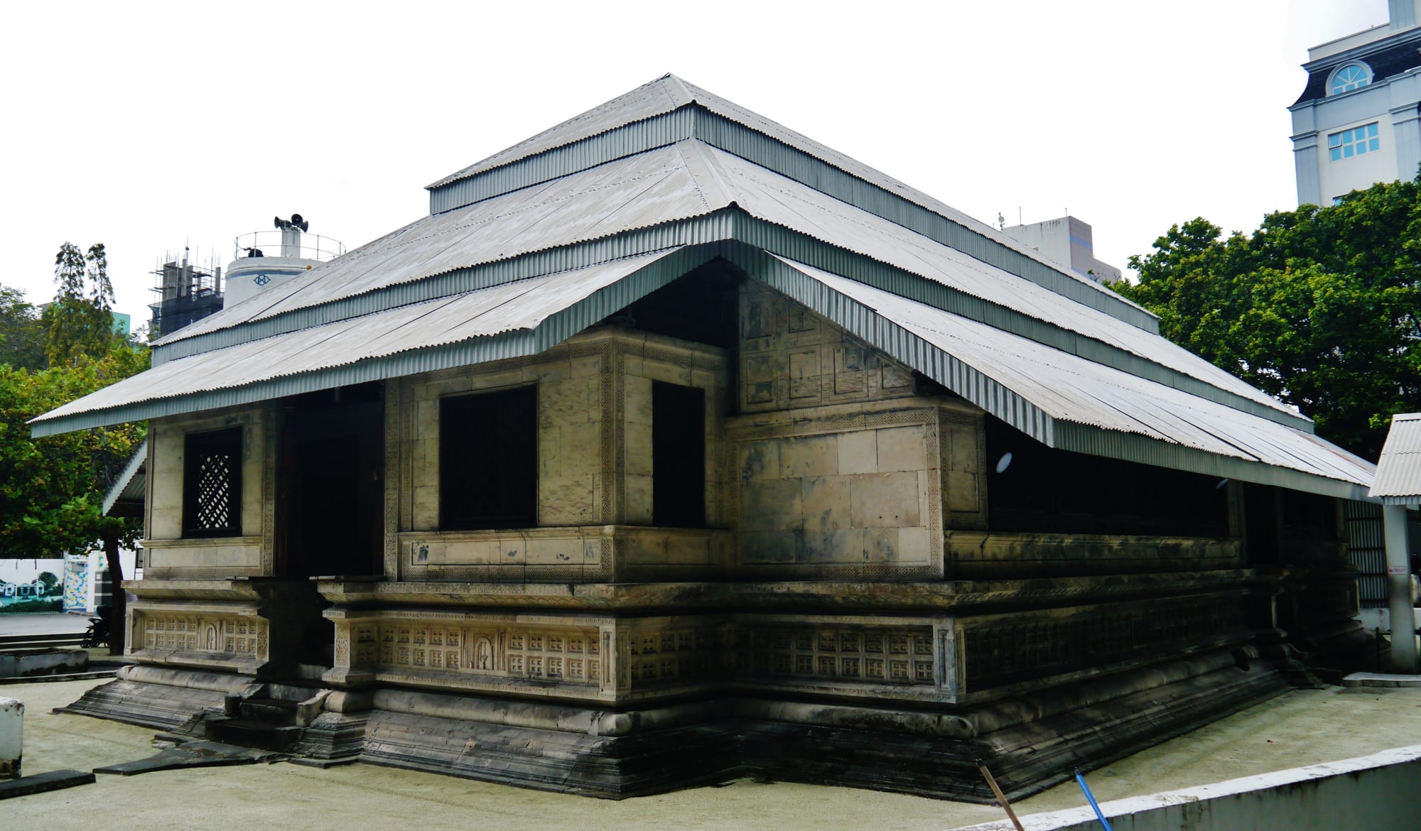 A low mosque of pale carved coral stone with lattice windows and decorated panels along its raised base, sheltered by a broad tiered metal roof, with trees and modern buildings behind