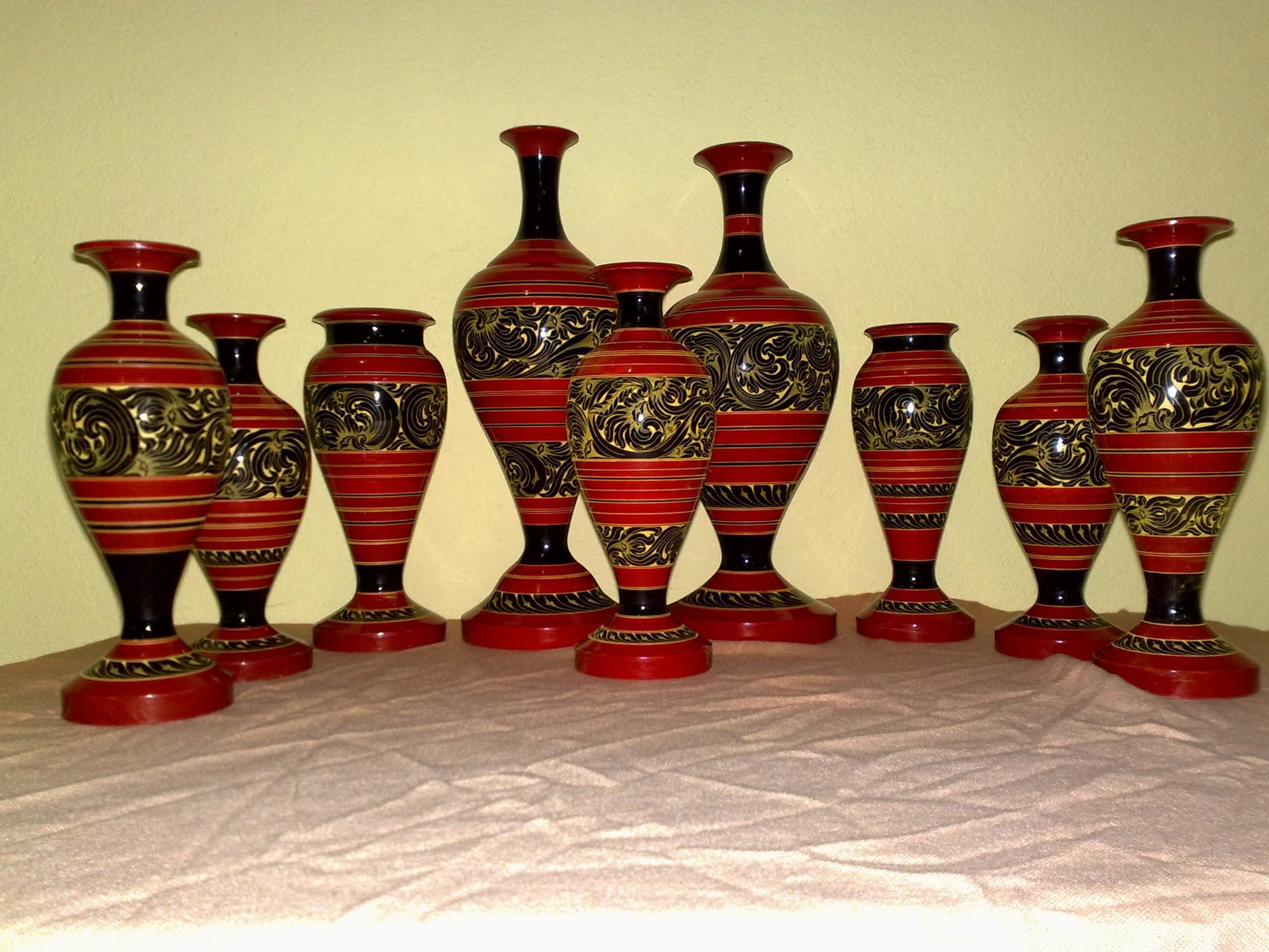 Nine turned wooden vases lacquered in bands of red and black, with swirling floral patterns in gold and black, lined up on a pale cloth