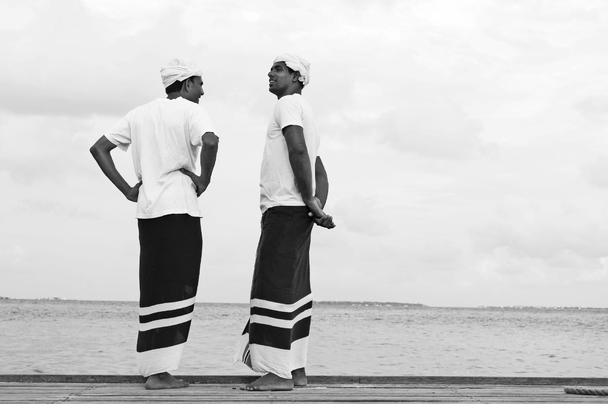 Black and white photograph of two barefoot men in white T-shirts and white headcloths, wearing long dark sarongs with white stripes at the hem, standing on a wooden jetty and talking, with the sea behind them