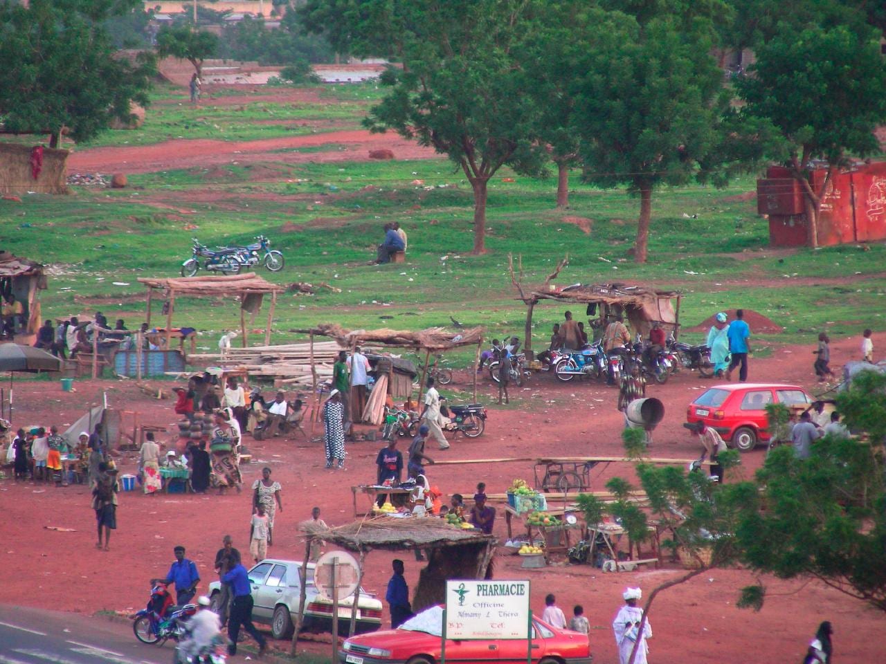A busy dirt-road market scene in Bamako with vendor stalls under thatched shelters, parked motorcycles, cars and pedestrians