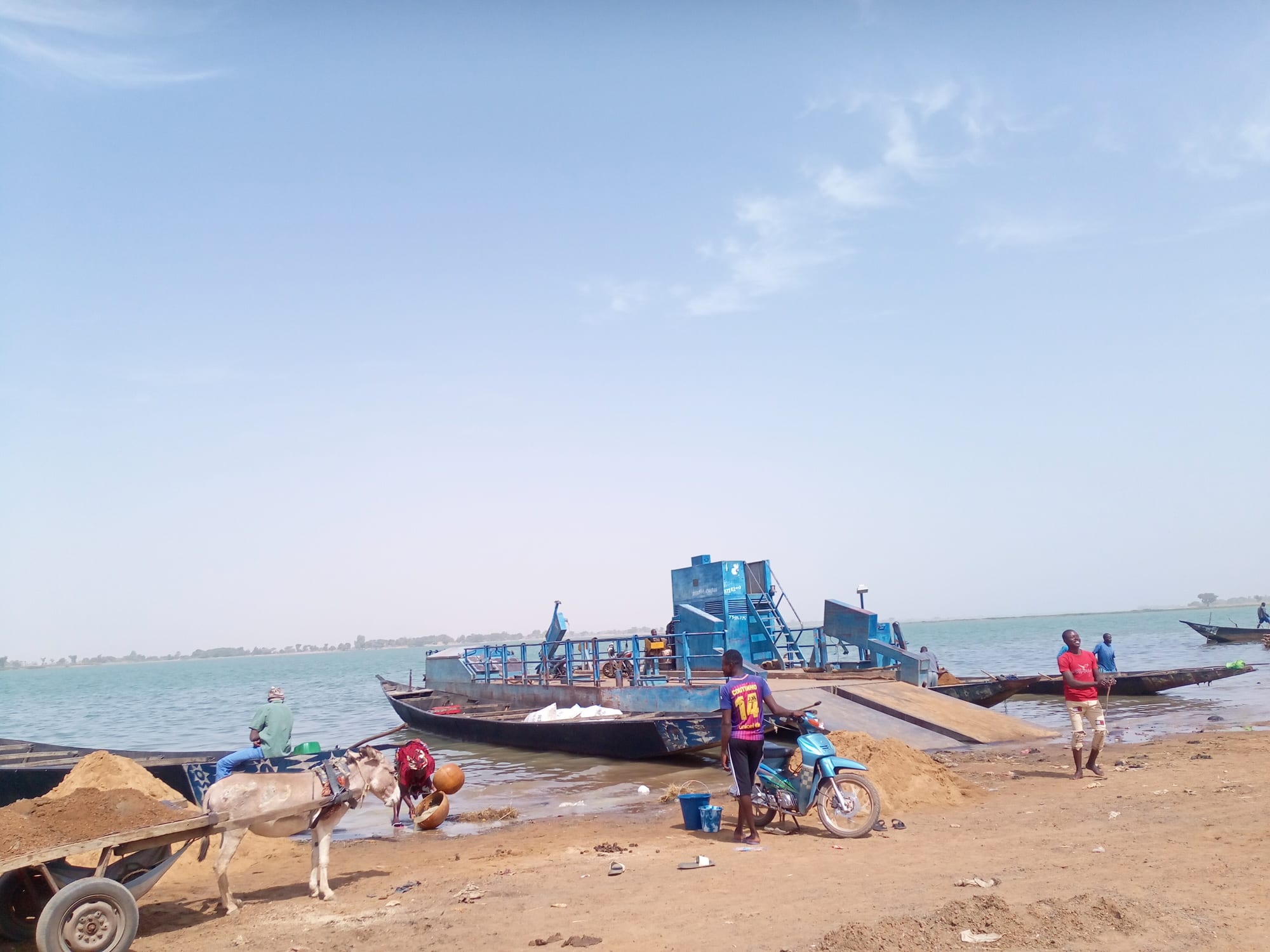 A motorised ferry and traditional wooden boats moored on the bank of the Niger River, with a donkey cart, a parked motorcycle and several people nearby