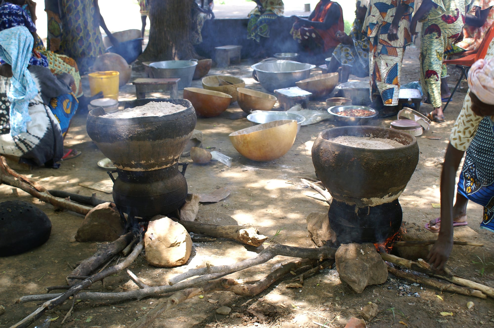 Women in colourful patterned clothing preparing millet in large blackened cooking pots set over open wood fires in a village courtyard