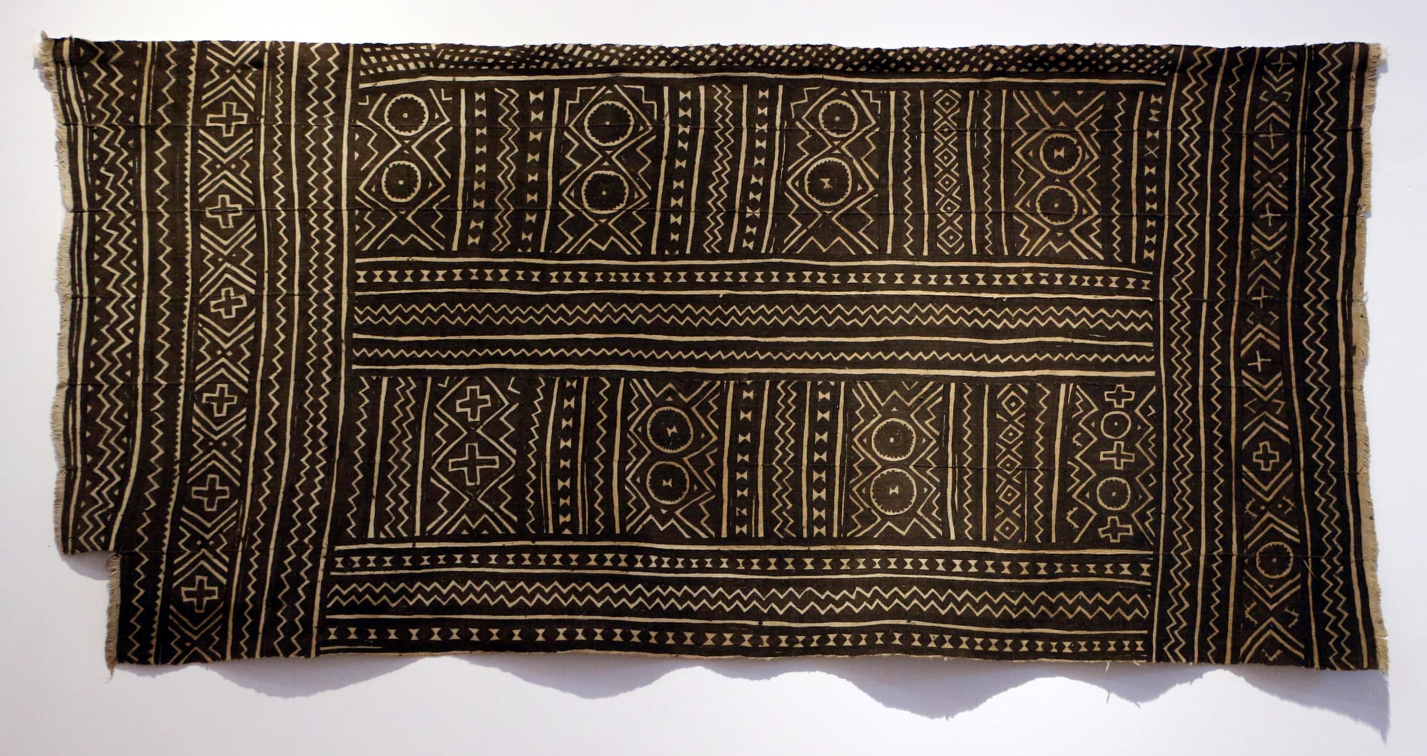 A dark brown bogolan cloth hung on a white wall, painted in cream with rows of zigzags, crosses, circles and diamond shapes