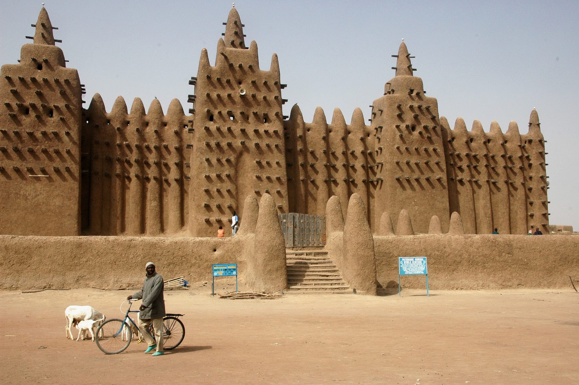 The Great Mosque of Djenné, a vast mud-brick building with three tall towers bristling with wooden beams, with a man wheeling a bicycle and two goats in the sandy square in front