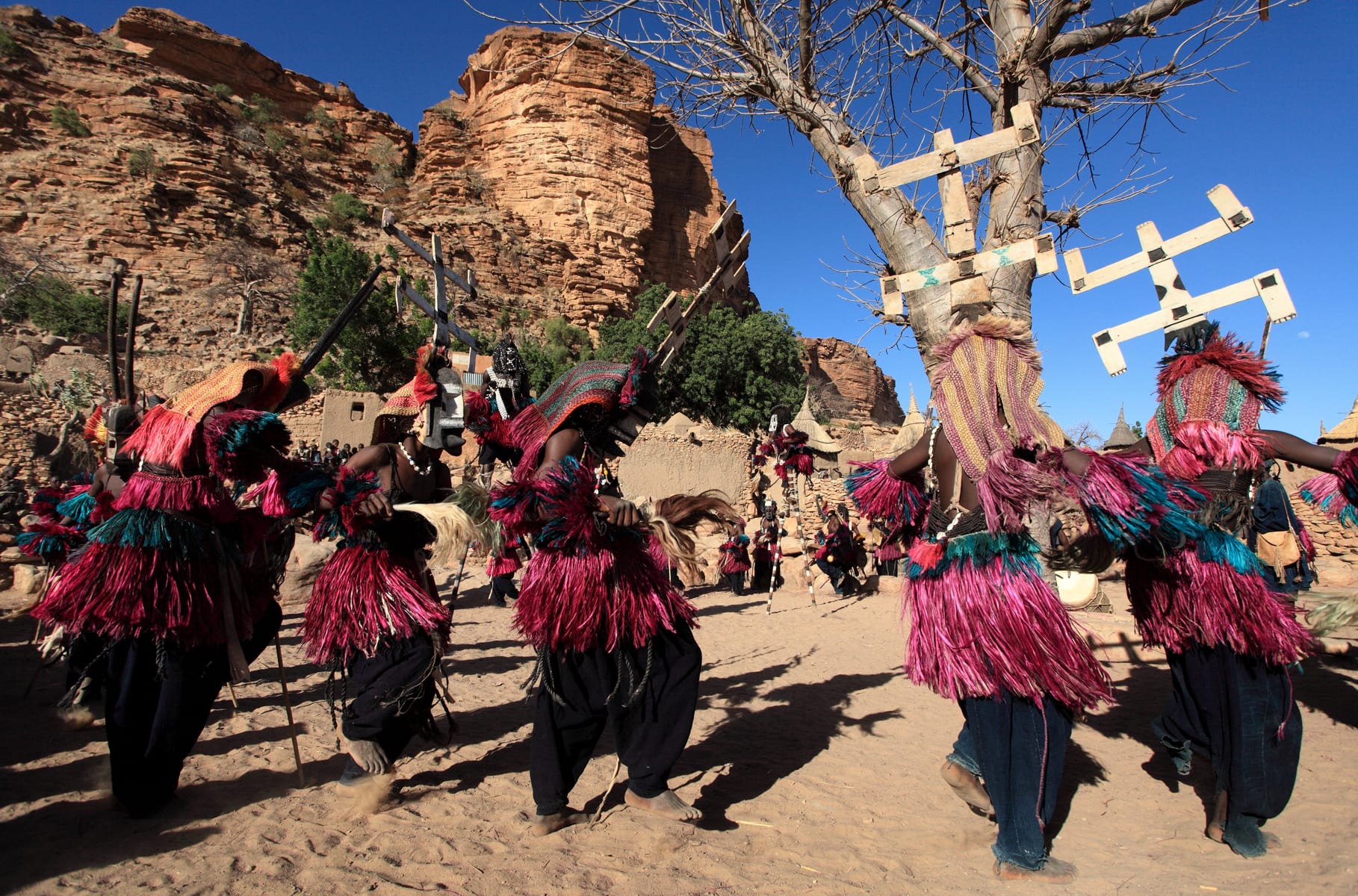 Masked Dogon dancers in red and pink fibre skirts and headdresses dance on sand beneath sandstone cliffs, several wearing tall masks topped with white double-barred crosses