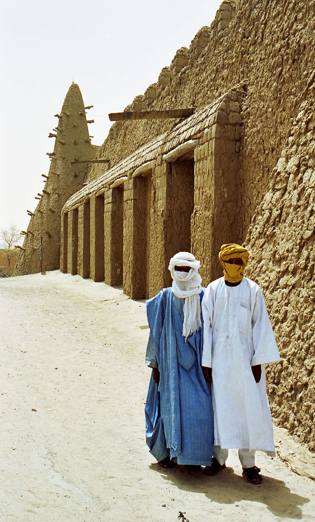A large mud-brick mosque with three pyramid-like towers standing in a sandy square
