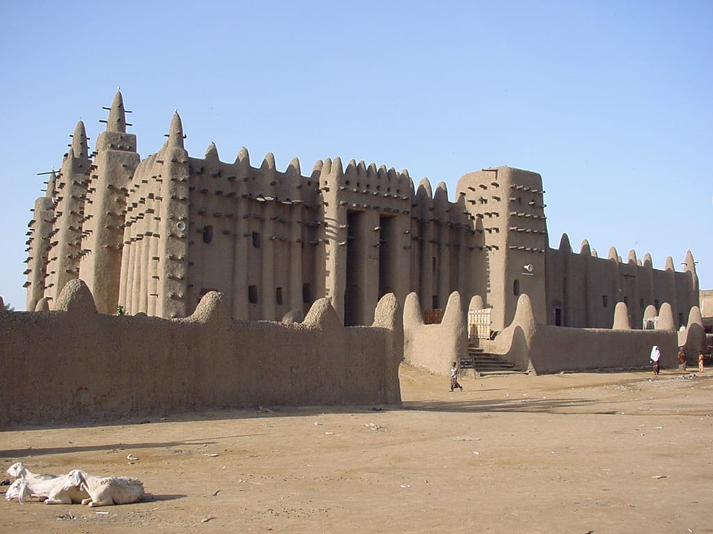 A vast mud-brick mosque with sculpted towers and protruding wooden beams, above a market square