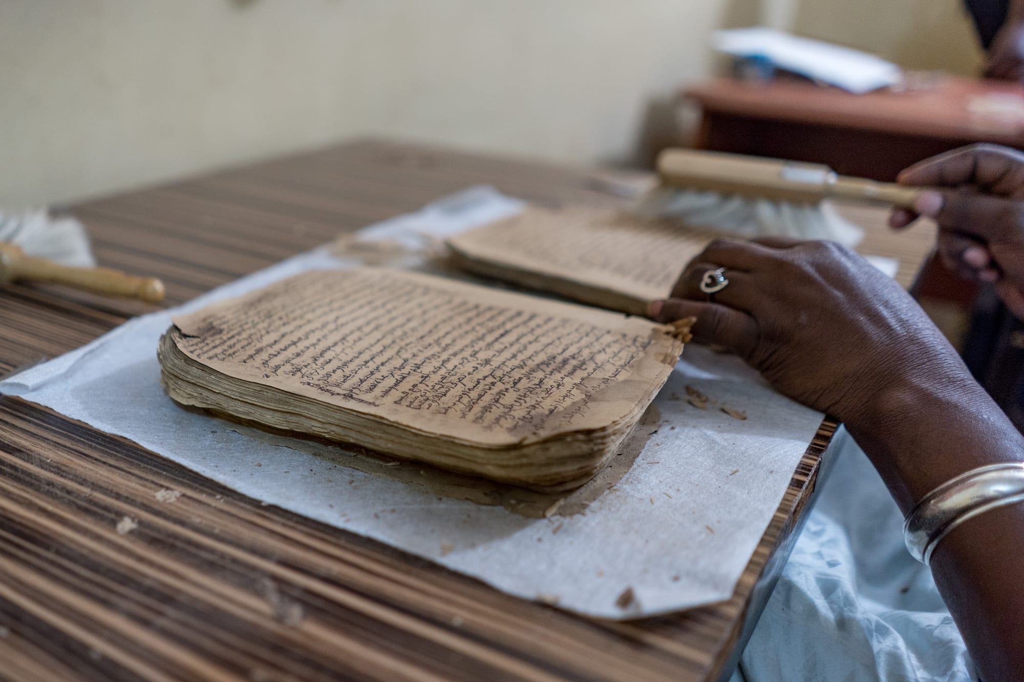 Hands resting beside a stack of loose, age-browned manuscript pages covered in handwritten Arabic script, with a soft brush on the table behind
