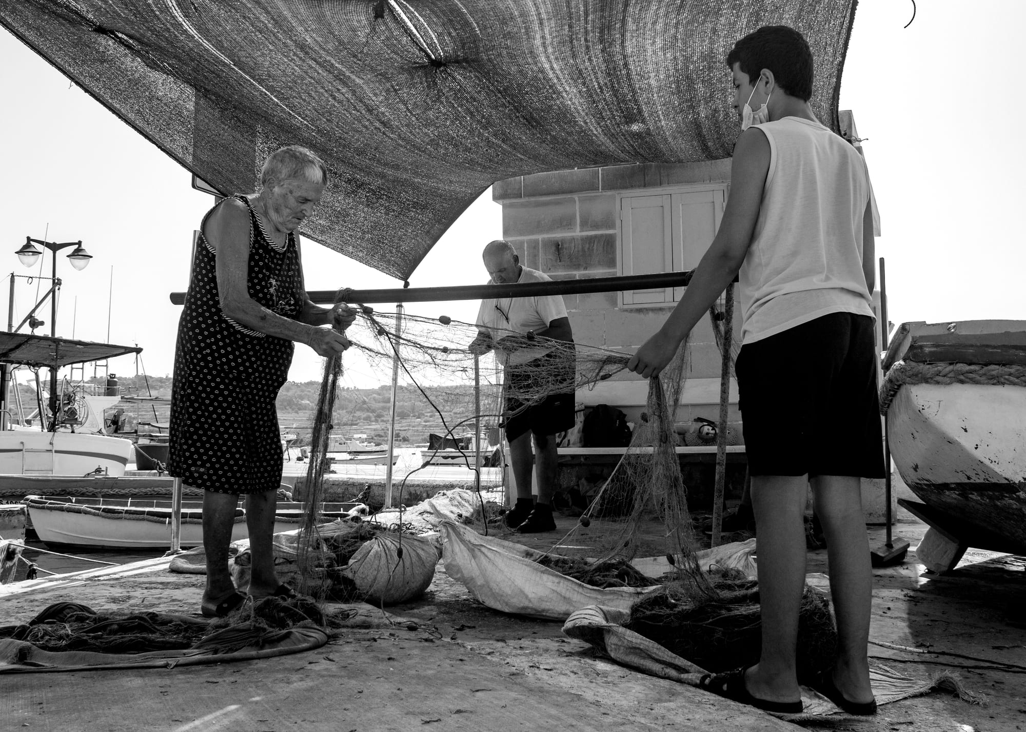 An elderly woman, a middle-aged man and a teenage boy work together cleaning and untangling a large fishing net under a shade awning at a harbourside