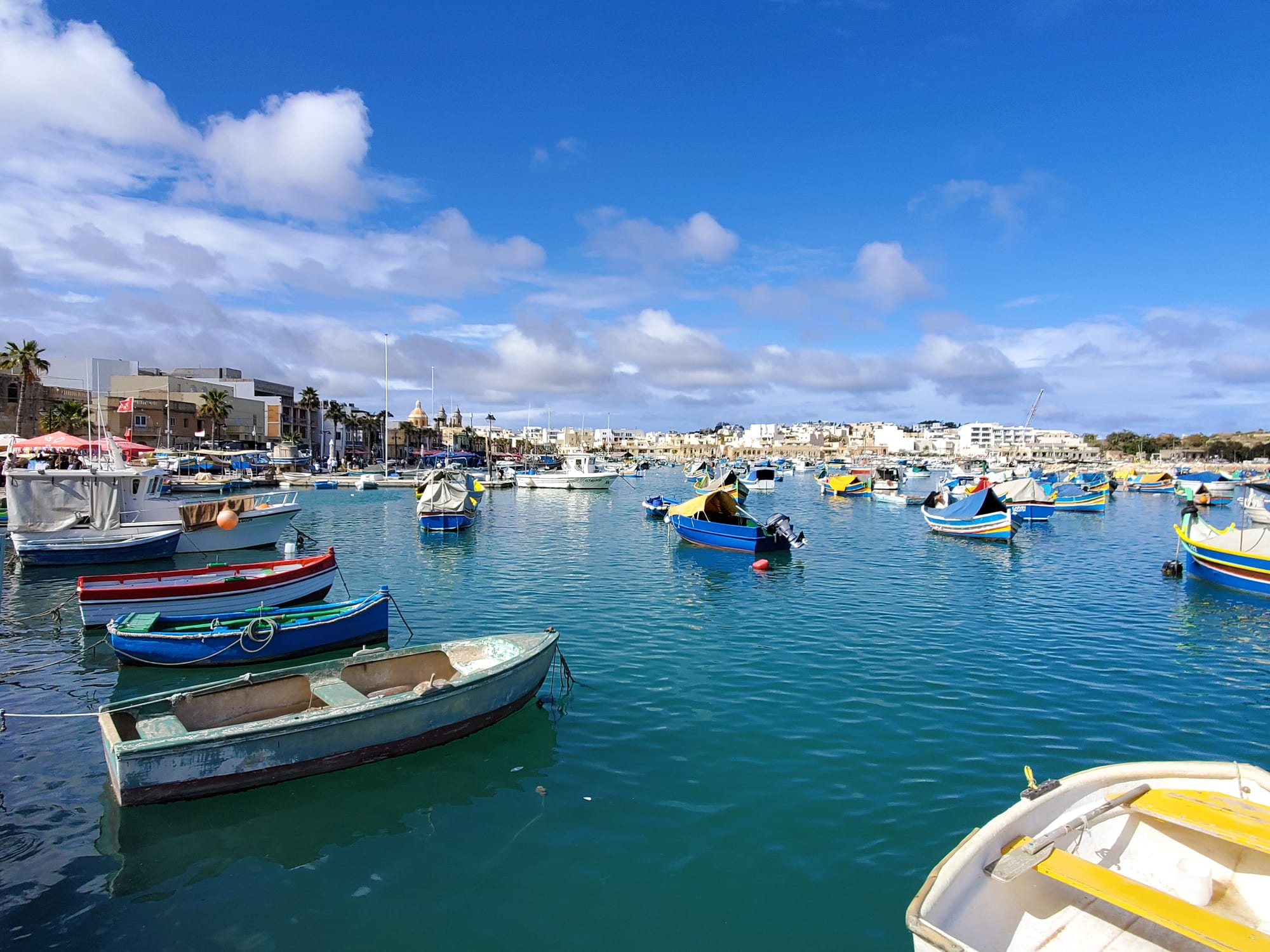 A harbour full of colourful traditional Maltese fishing boats called luzzus, moored in clear turquoise water with a village and market stalls visible on the shore