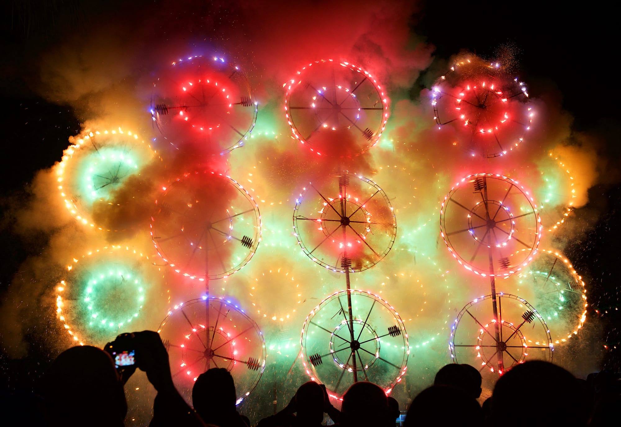 A night-time display of large illuminated wheels spinning and throwing out red, green and white sparks amid clouds of coloured smoke, with the silhouetted heads of spectators and a raised phone camera in the foreground