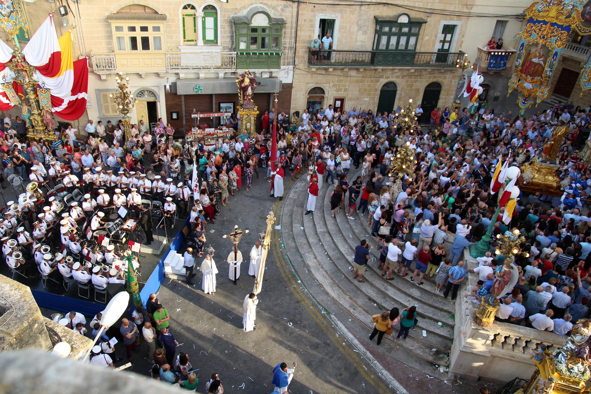 Seen from above, a marching band in white caps plays in a crowded village square as robed clergy and altar servers carry a crucifix and processional staffs, with decorated statues on pedestals and festa banners around the square