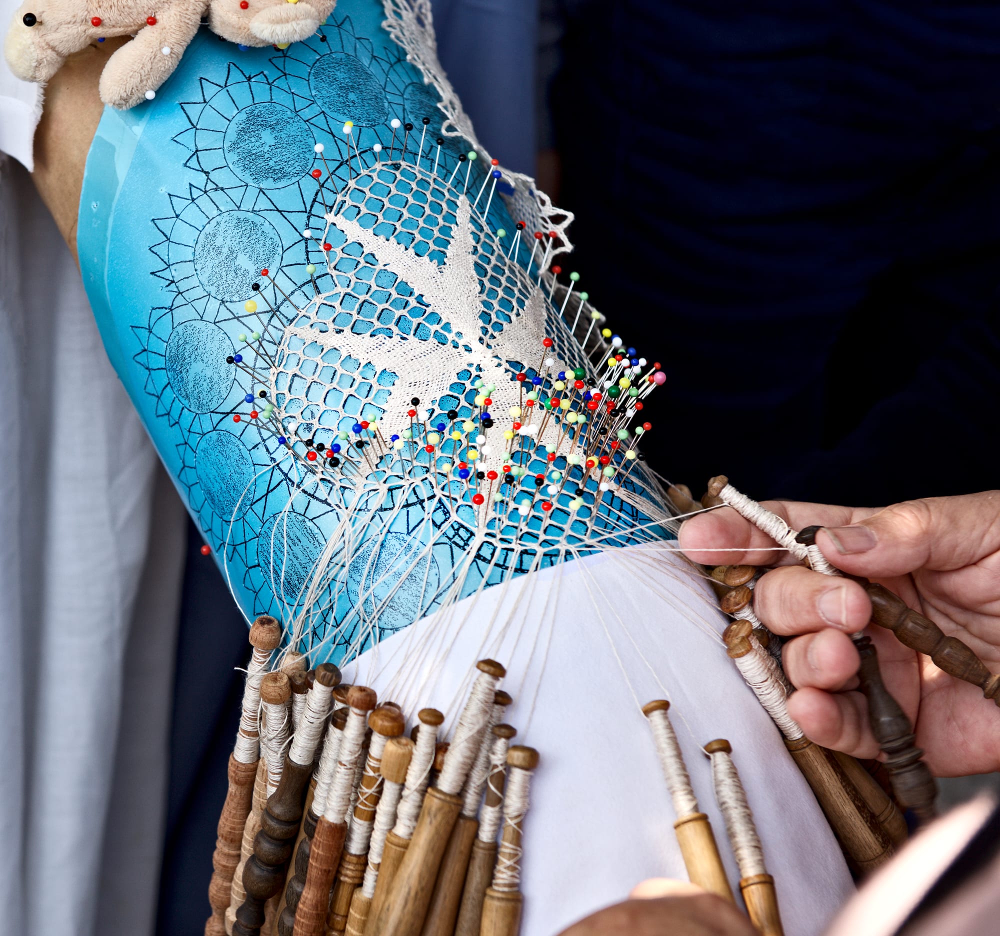 Close-up of a lace pillow covered in coloured pins, with a white eight-pointed cross taking shape in bobbin lace and dozens of wooden bobbins hanging below, one hand working the bobbins