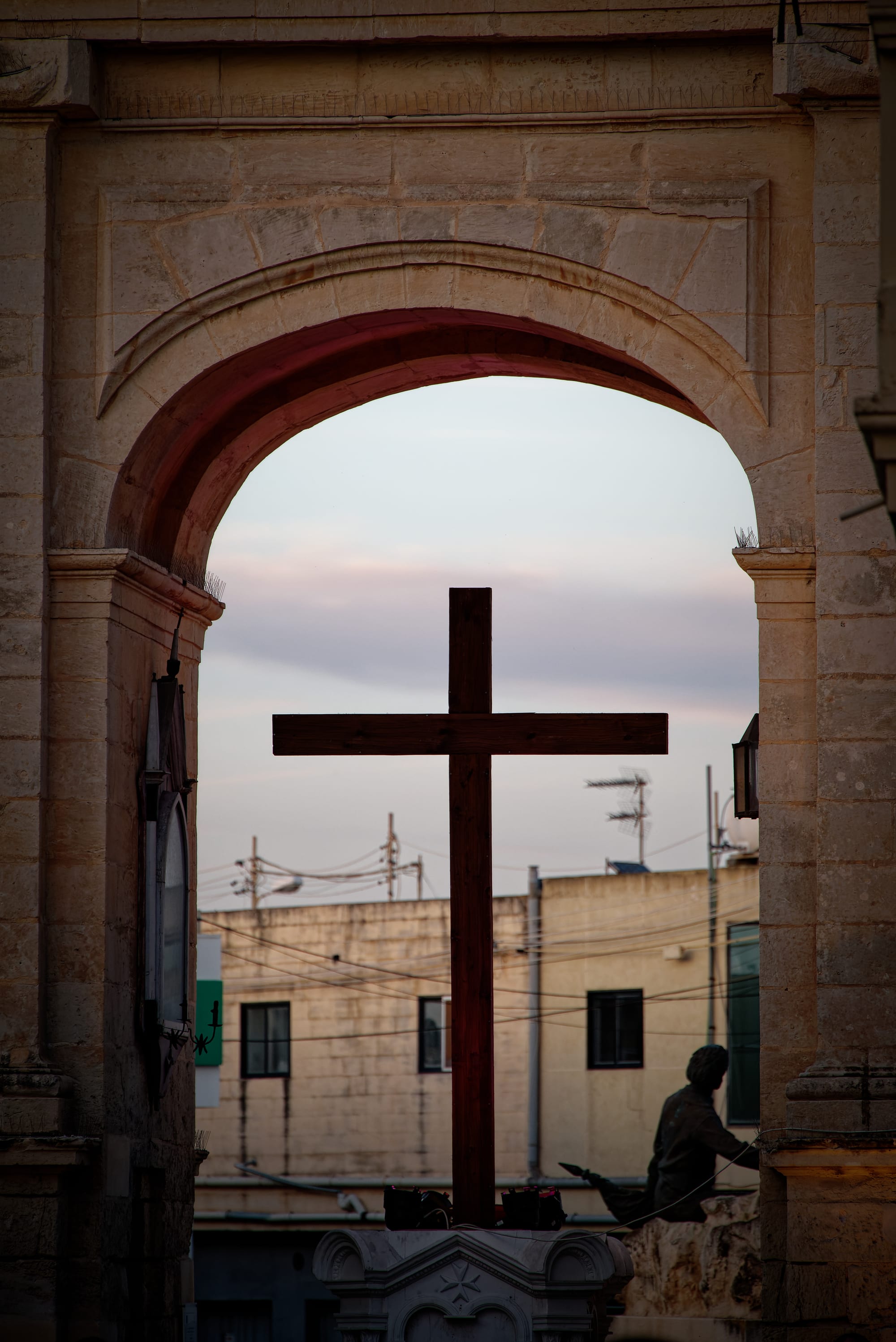 A plain dark wooden cross silhouetted against a pale evening sky, framed by a tall stone archway, with buildings and overhead wires behind it