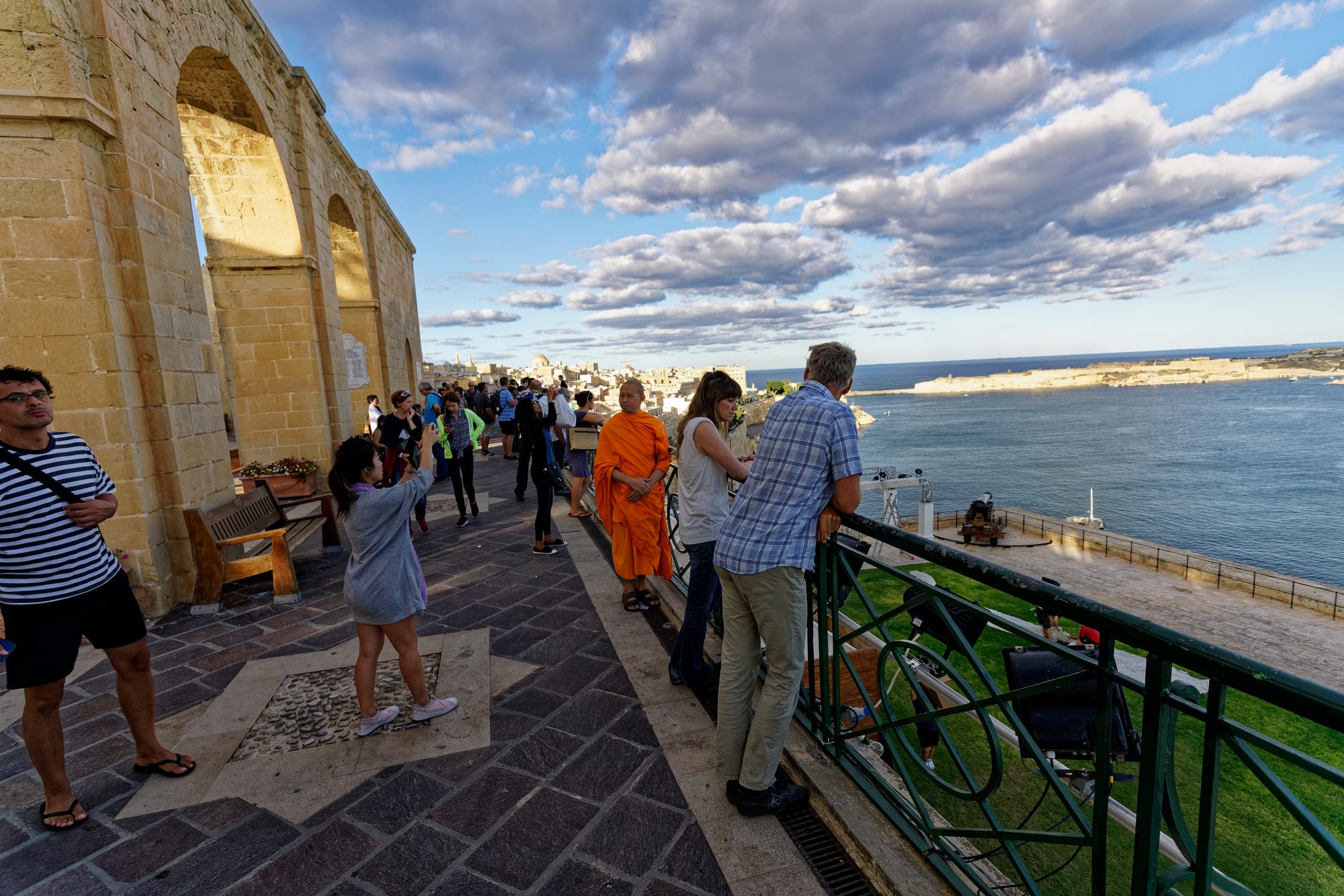 A deep natural harbour ringed by golden-stone fortifications and church domes, seen from a garden terrace