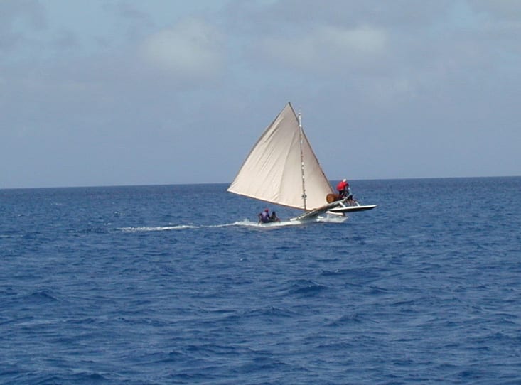 A traditional outrigger sailing canoe under a triangular sail, cutting through open ocean