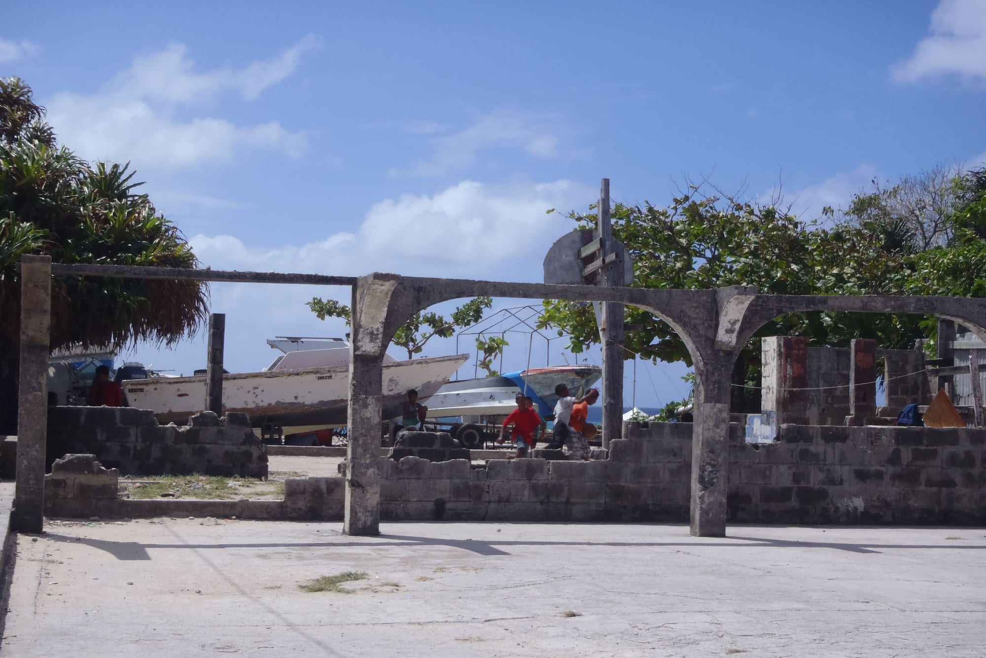 Children playing basketball on a concrete court squeezed between boats and cinderblock walls in Ebeye, with a hoop mounted on a concrete frame