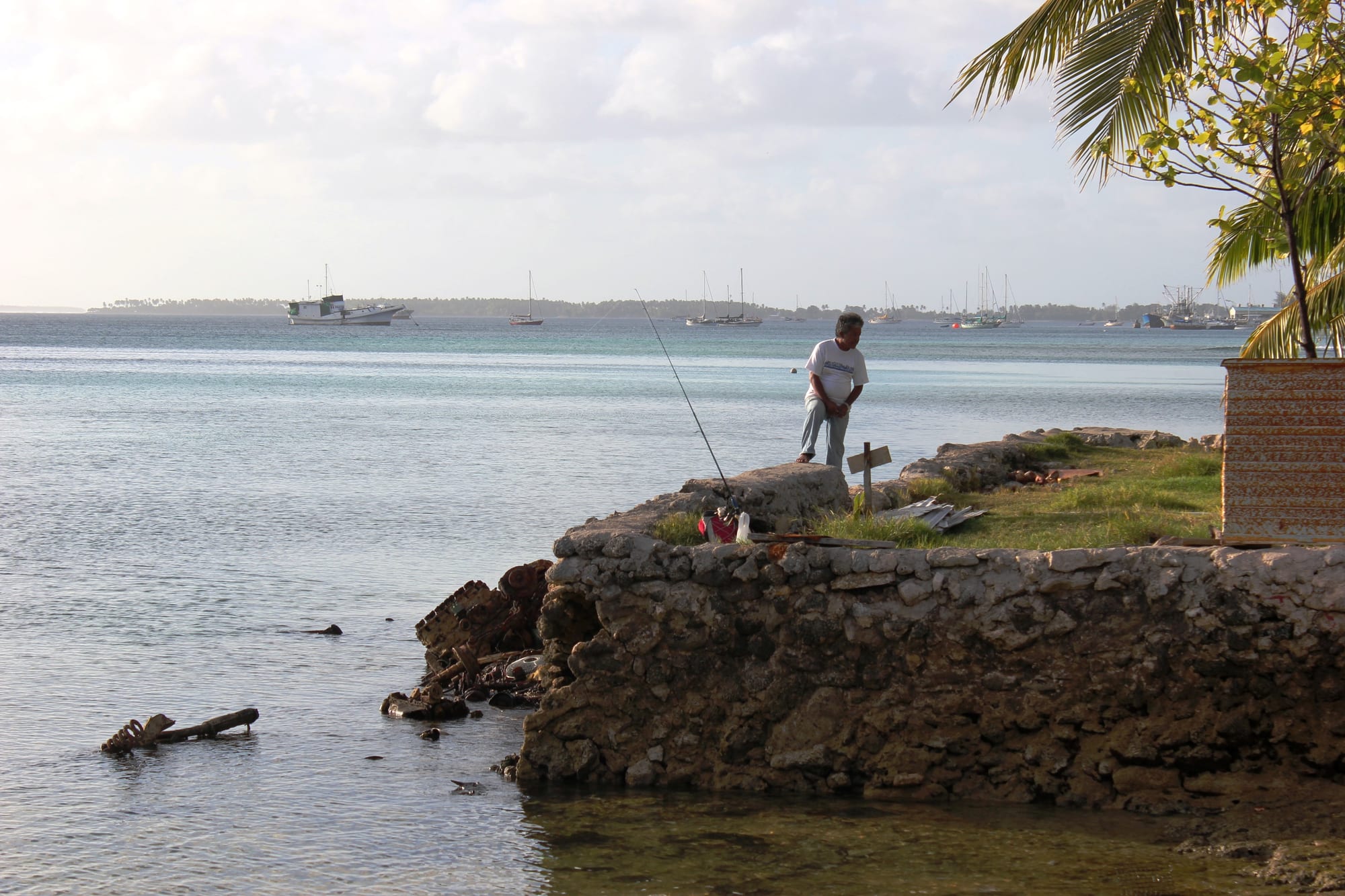 A man fishes with a rod from a low stone seawall beside a calm lagoon, with several boats moored in the water behind him