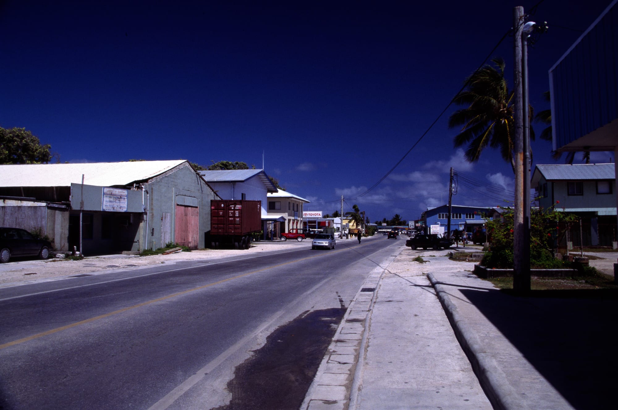 A quiet single paved road running through the small town centre of Majuro, lined with low corrugated-metal shops and a few parked cars